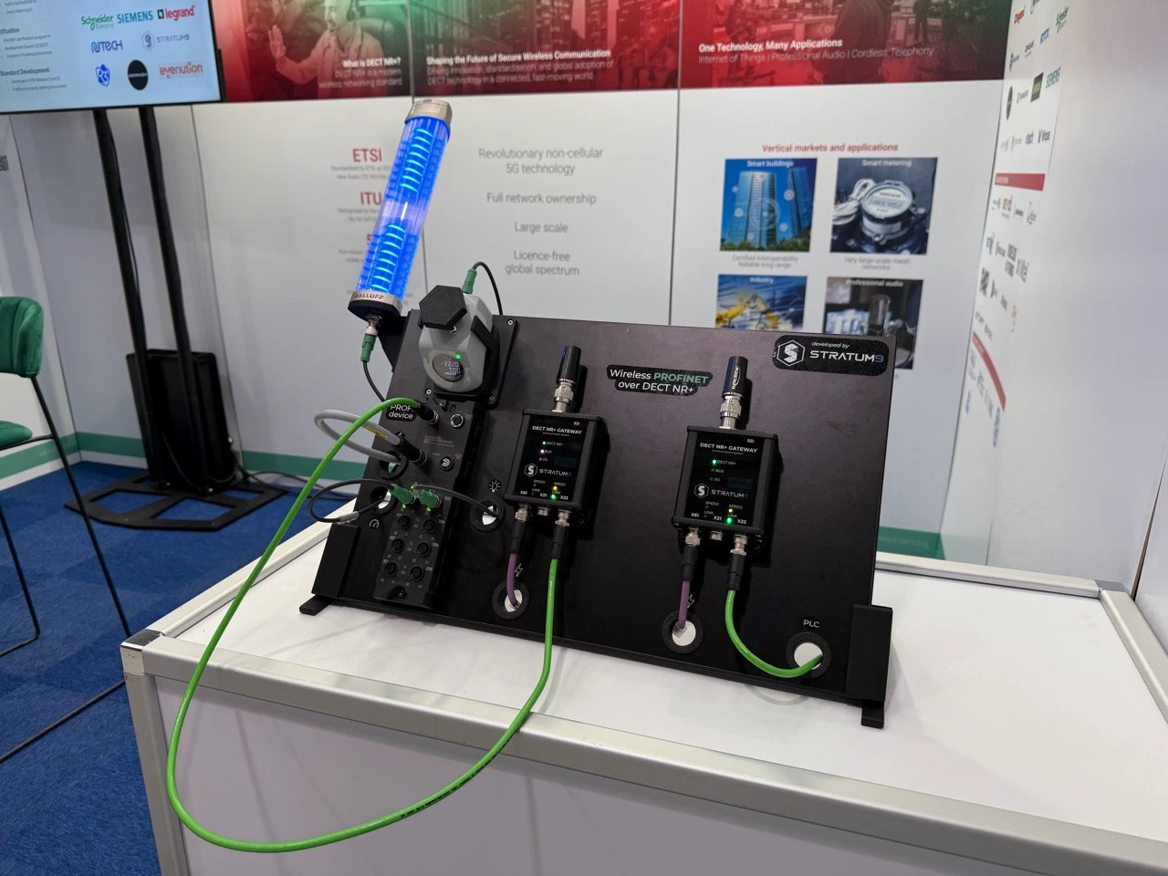 Demo-Board „Wireless PROFINET over DECT NR+“ von STRATUM 9 auf einem Messestand. Zwei DECT NR+ Gateways mit Antennen verbinden die Steuerung (PLC) drahtlos mit einem PROFINET-IO-Modul. Am Modul hängen eine blau leuchtende Signalsäule und ein Sensor mit Display.