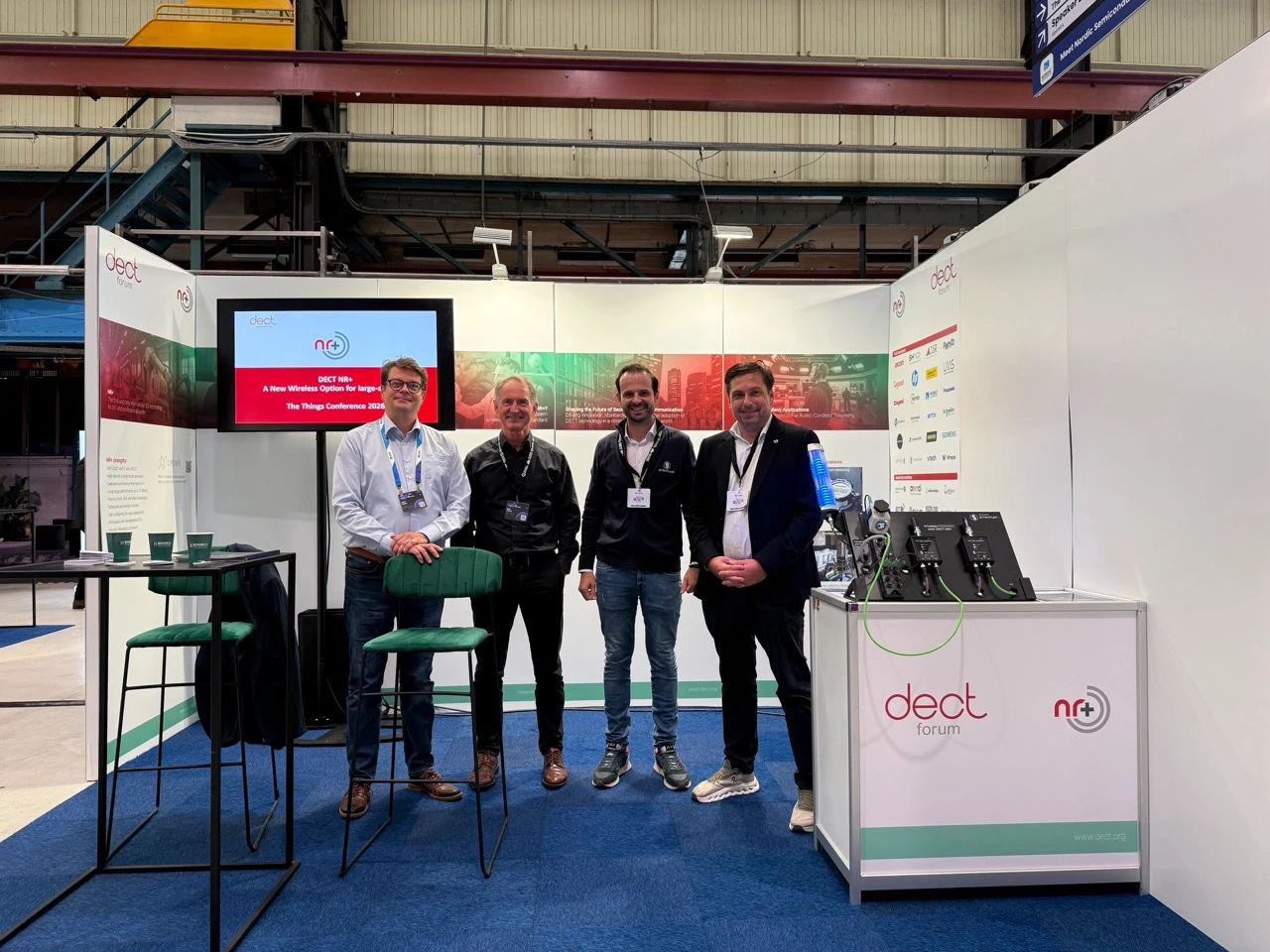 Vier Männer stehen nebeneinander am Stand des DECT Forums auf der Things Conference 2026 in Amsterdam. Von links: Lauri Piikivi, Roel Ottink, Alfred della Torre Jr. von STRATUM 9 und Christoph Gulich. Hinter ihnen zeigt ein Bildschirm eine Folie zu DECT NR+. Rechts steht auf dem Standtresen das Demo-Board „Wireless PROFINET over DECT NR+“ von STRATUM 9.
